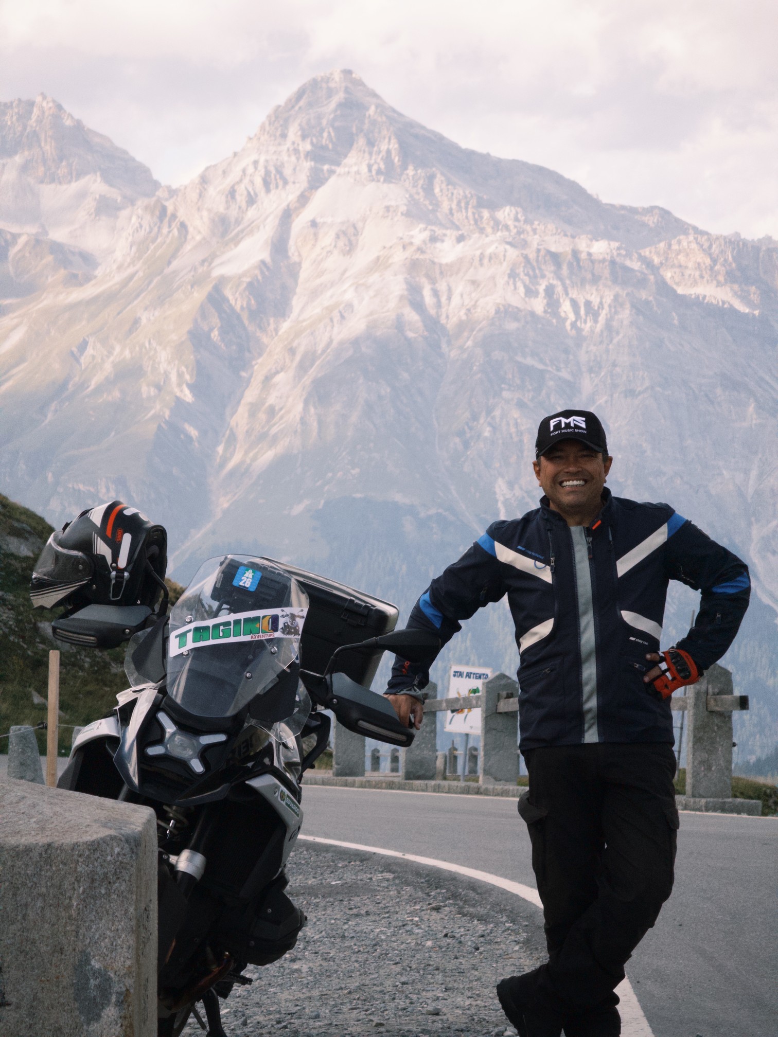 João Tagino sorrindo ao lado da moto, numa estrada de montanha